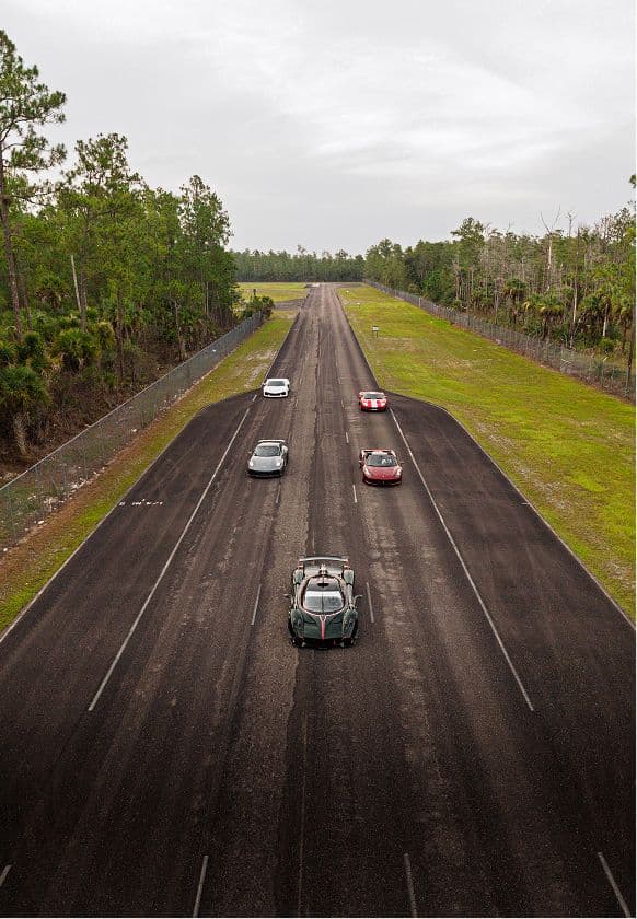 Five sports cars staged across the three-lane straightaway, seen from above, with pine woodland on either side.